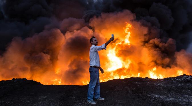 Seorang pria berselfie di depan kobaran api kilang minyak saat operasi perebutan kota Mosul oleh Pasukan Irak dari tangan ISIS, di Qayyarah, Mosul (19/10). Militer Irak telah berhasil menguasai beberapa wilayah ISIS di Mosul. (AFP Photo/YASIN Akgul)